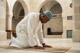 Homme en djellaba blanche effectuant la prière dans une mosquée de Grenoble avec mihrab visible en arrière-plan